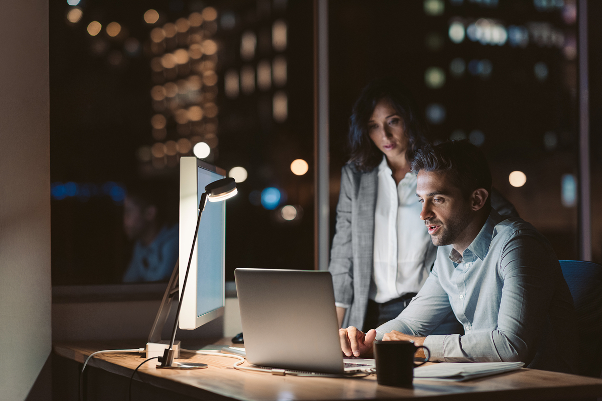 Woman standing over man's shoulder while he sits at a desk. Both are looking at the laptop screen.