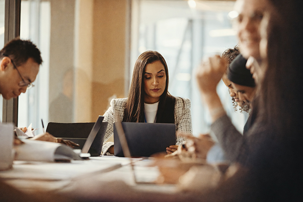 A group of people sitting around a meeting table working on their laptops and writing notes.