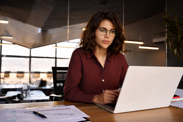 A woman wearing glasses and red shirt sitting at a desk looking at a computer