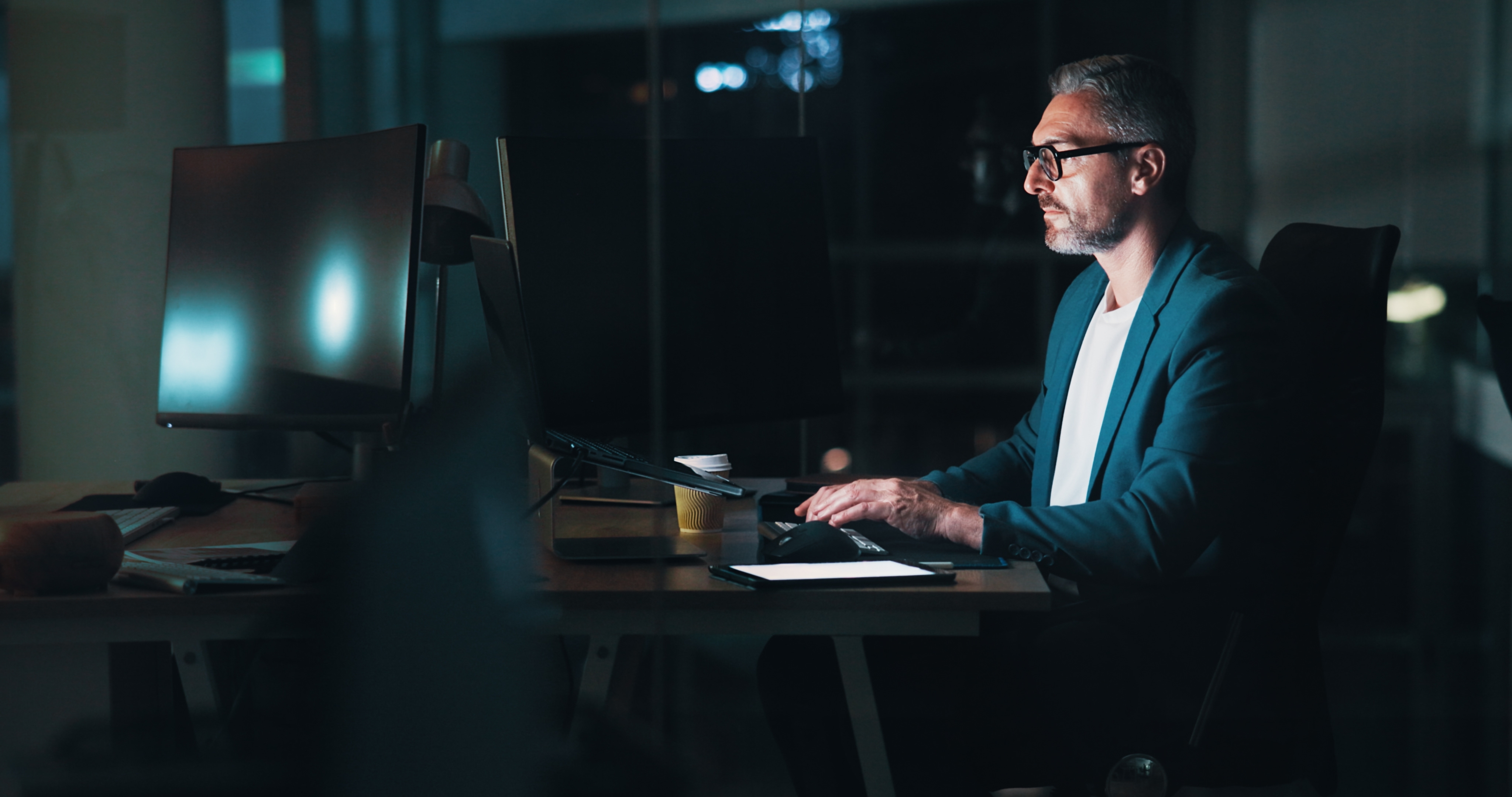 Man wearing glasses sitting at a desk working on a computer late at night