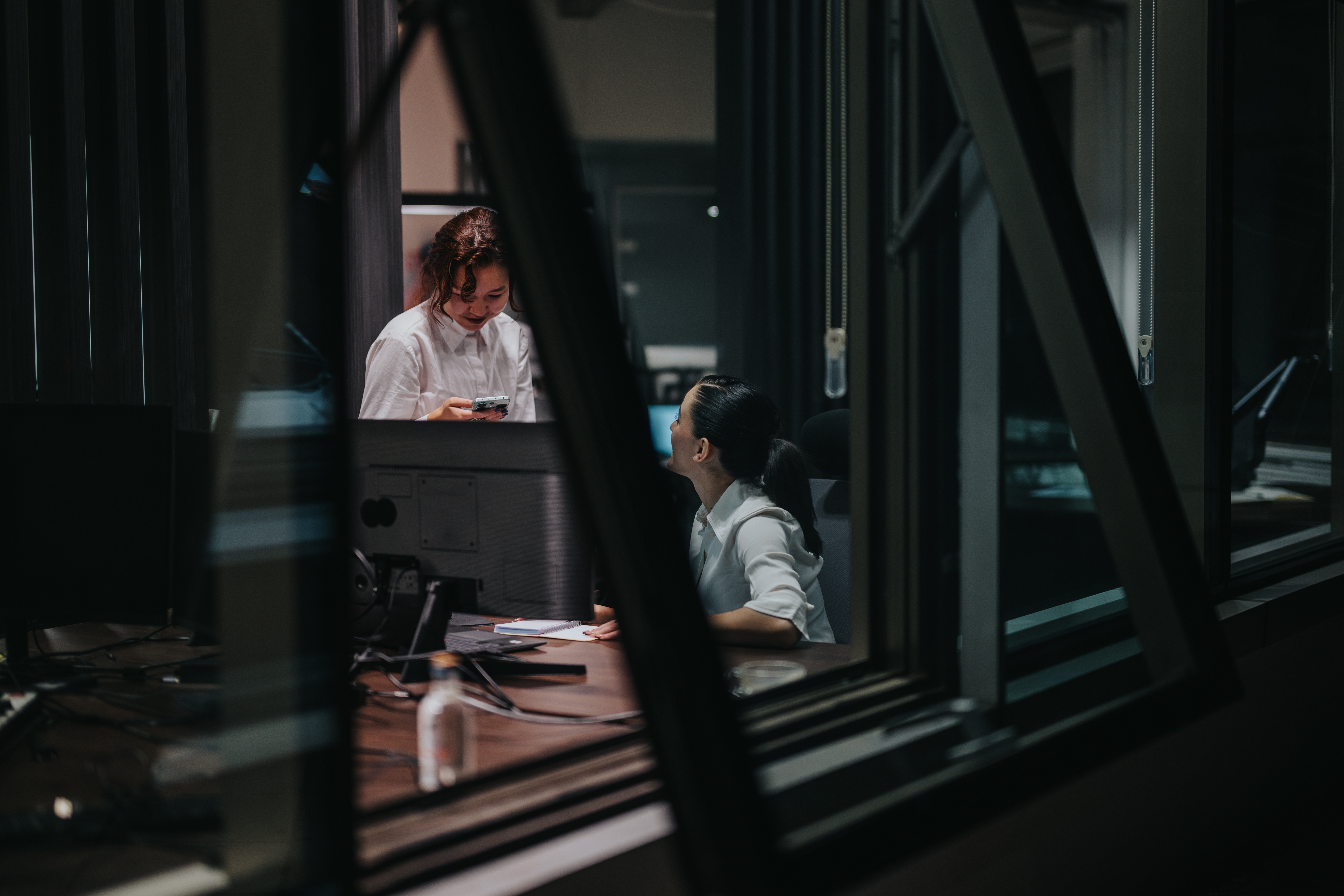 Candid shot of two coworkers talking to each other at a desk late at night